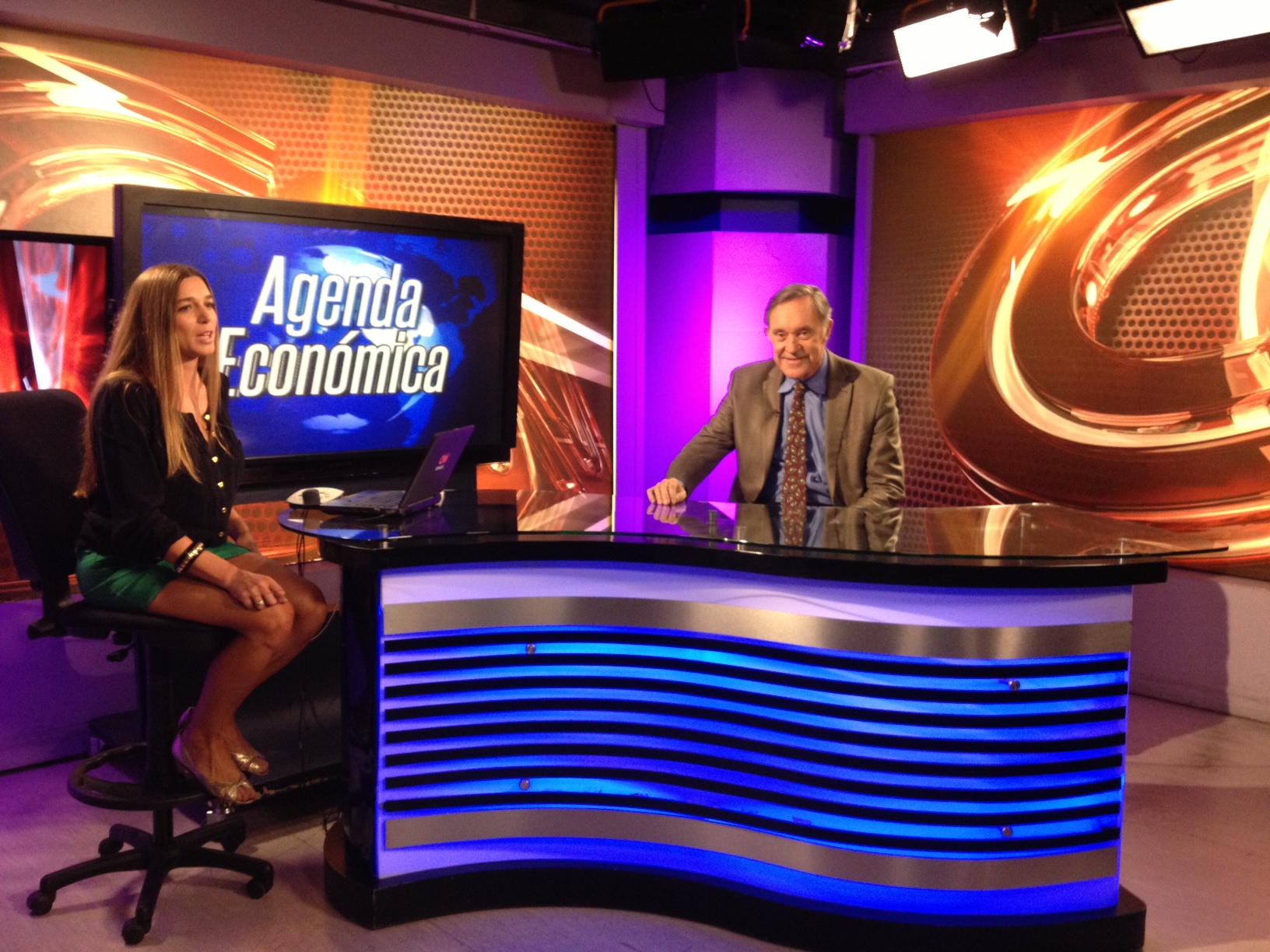 A male guest and female host sit on a vibrant news set labeled "Agenda Económica," both ready for a televised discussion.