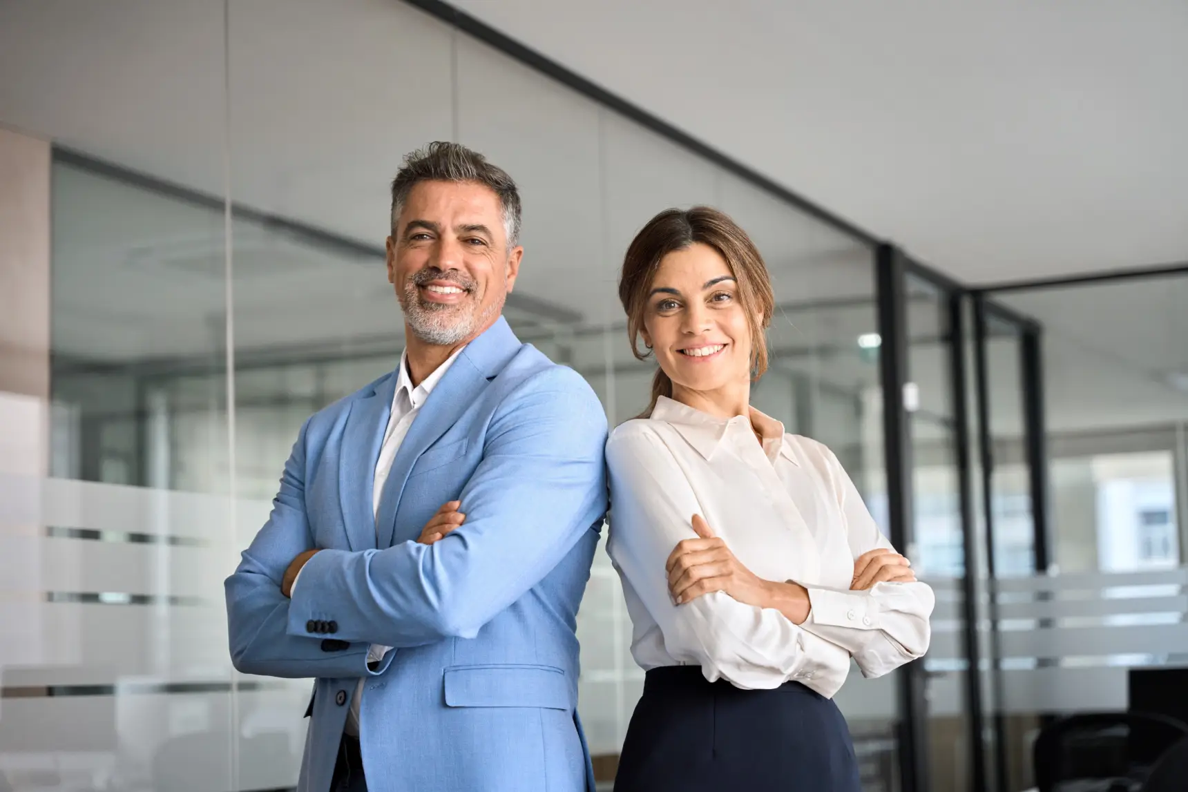 Two happy executive managers, mature business man and woman leaders, successful partners professional team in their 50s standing arms crossed in office at work looking at camera. Corporate portrait.